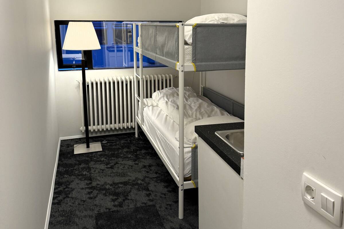 Habitación de hotel compacta con literas, un radiador, una ventana con lámpara y un lavabo.
