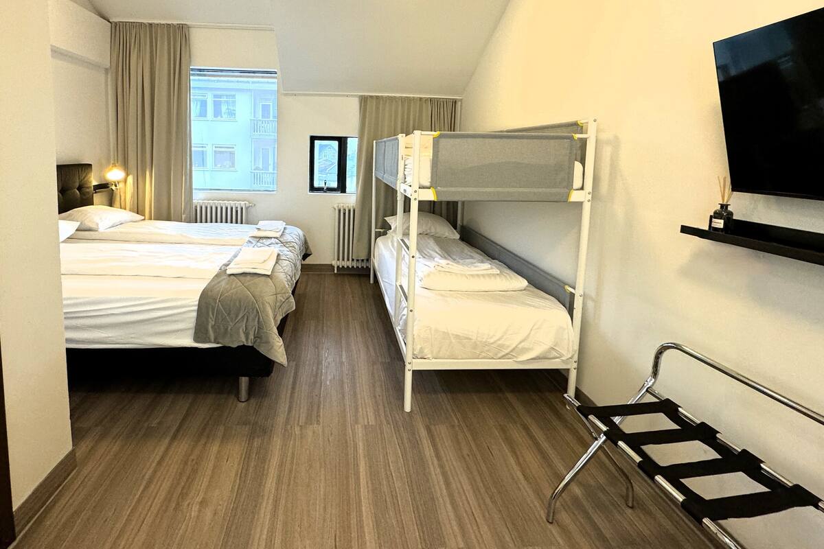 Habitación de hotel con dos camas, una litera, un radiador, una ventana con cortinas, un televisor montado en la pared y un estante pequeño con una botella.