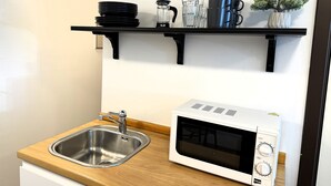 Una cocina con un lavamanos de acero inoxidable, un microondas y estantes negros con platos, vasos y una cafetera.