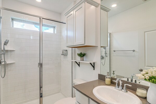 Un baño moderno con ducha acristalada, azulejos blancos, encimera beige, lavabo con espejo y una pequeña estantería con una planta.