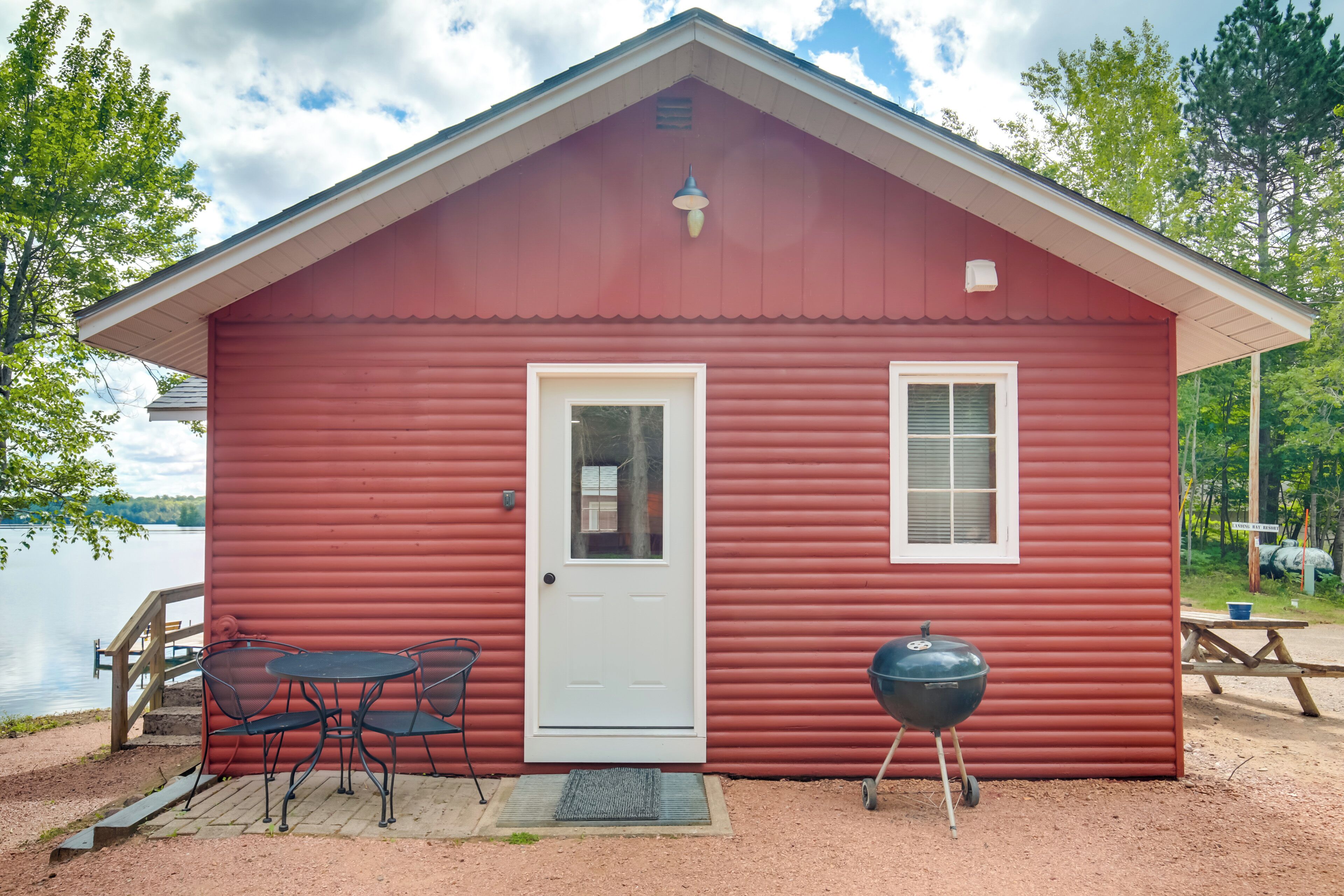 Une cabane rouge avec une porte blanche, un petit porche avec une table et des chaises, et un barbecue à l’extérieur.