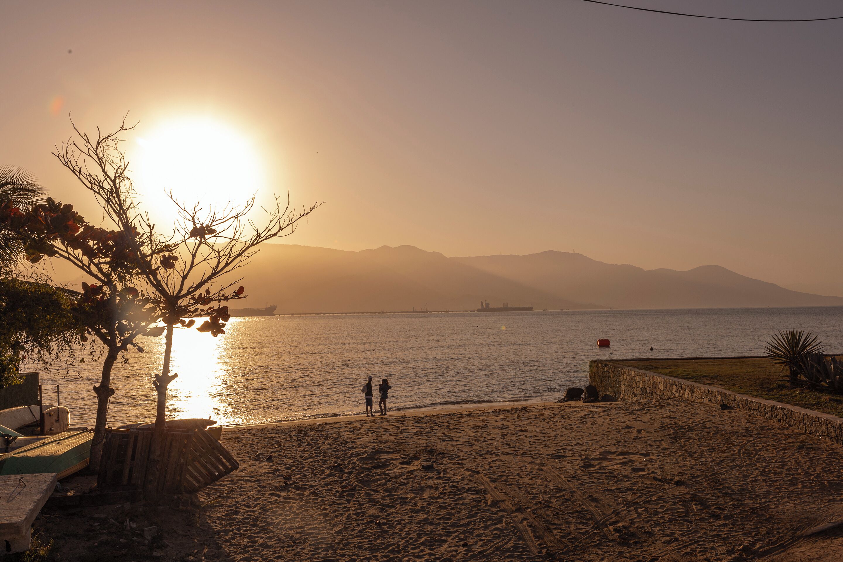A beach with boats, a stone wall, and two people walking near the water.