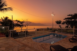Zona de piscina con vista al atardecer sobre el agua, rodeada de palmeras y un patio de piedra.