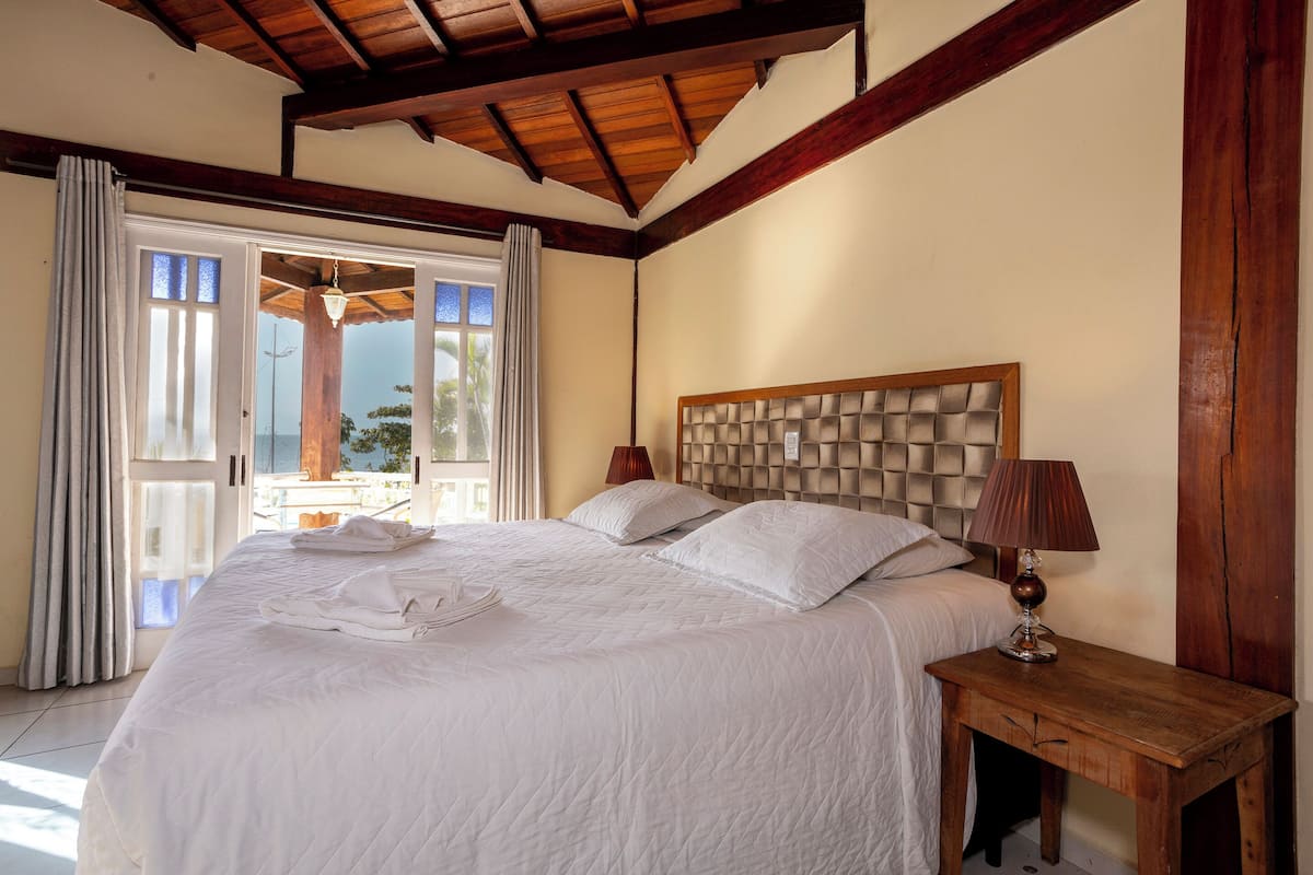 A neatly made bed with a wooden headboard, a bedside table with a lamp, and a view through a glass door to the outside.
