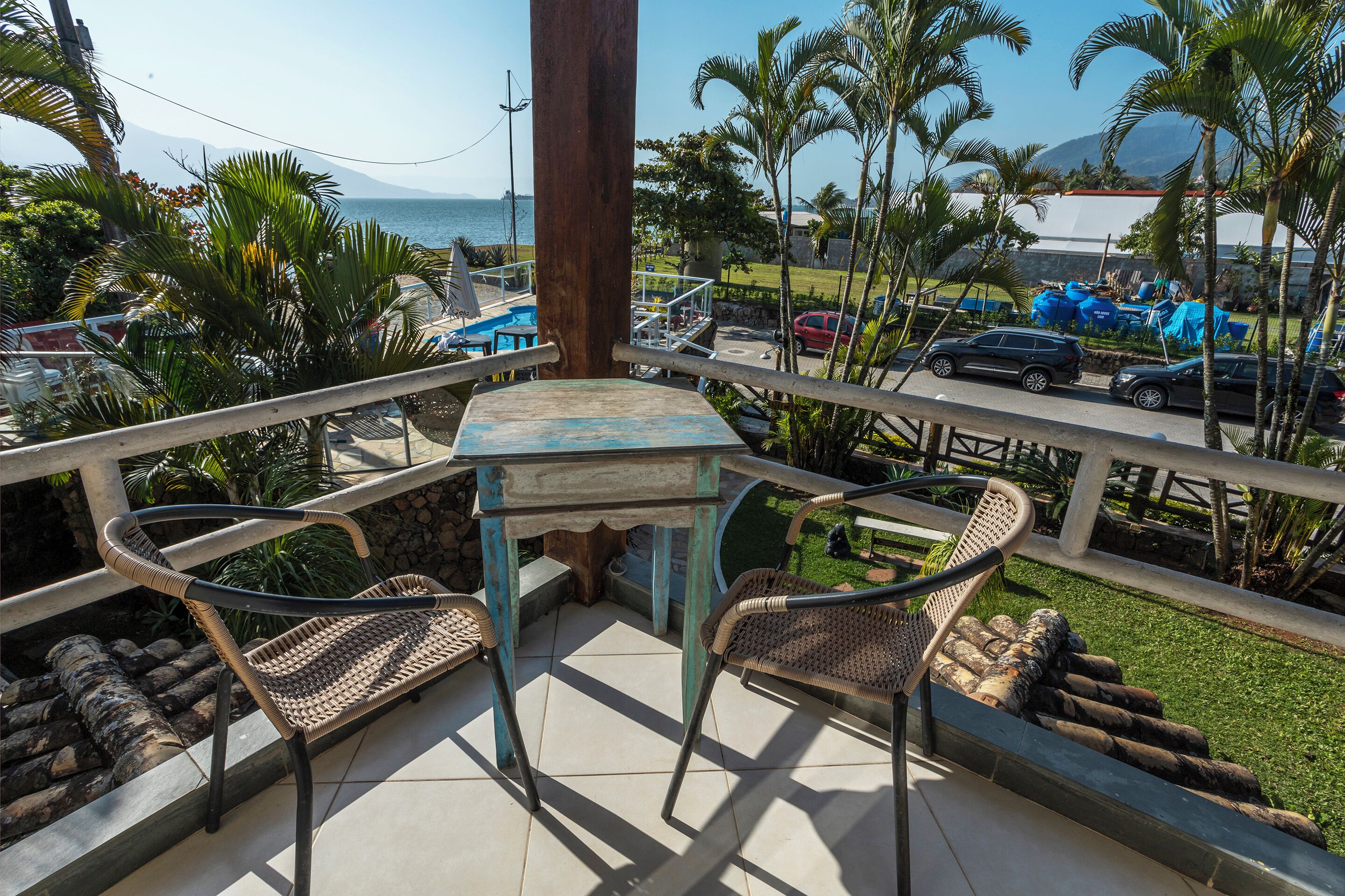 A balcony with a table and chairs, overlooking a pool and a parking area with cars.