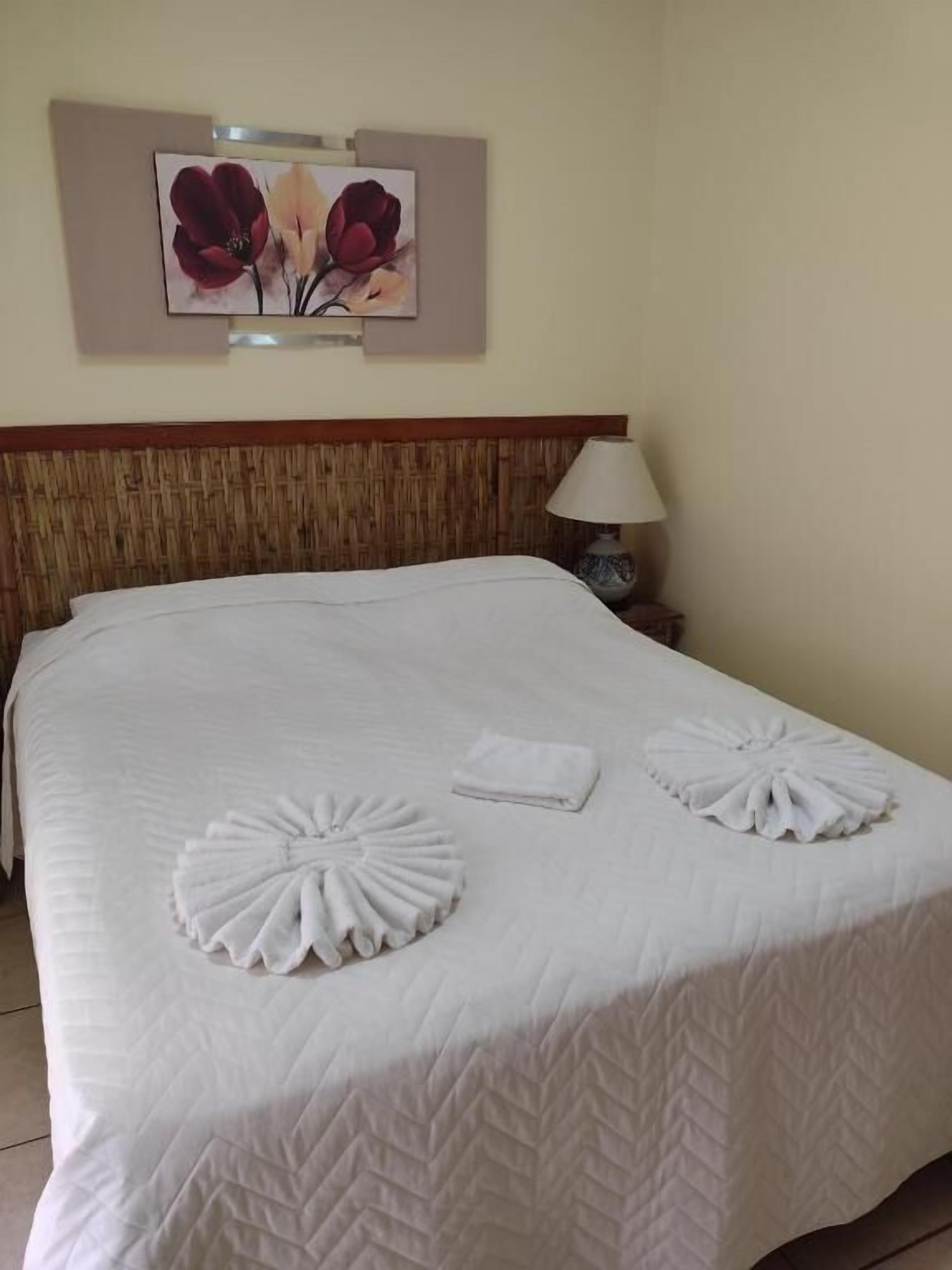 A neatly made bed with a white quilt and two towel turkeys, a wooden headboard, a wall-mounted lamp, and a framed floral artwork above the bed.