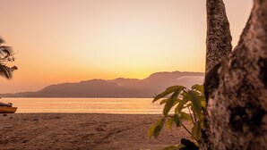 A beach with a boat, palm trees, and mountains in the background. - Pousada Bonns Ventos (Ilhabela)
