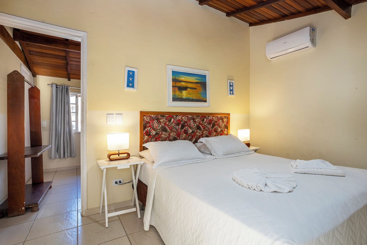 A hotel room with a bed, two bedside lamps, a painting on the wall, an air conditioner, and a wooden ceiling.