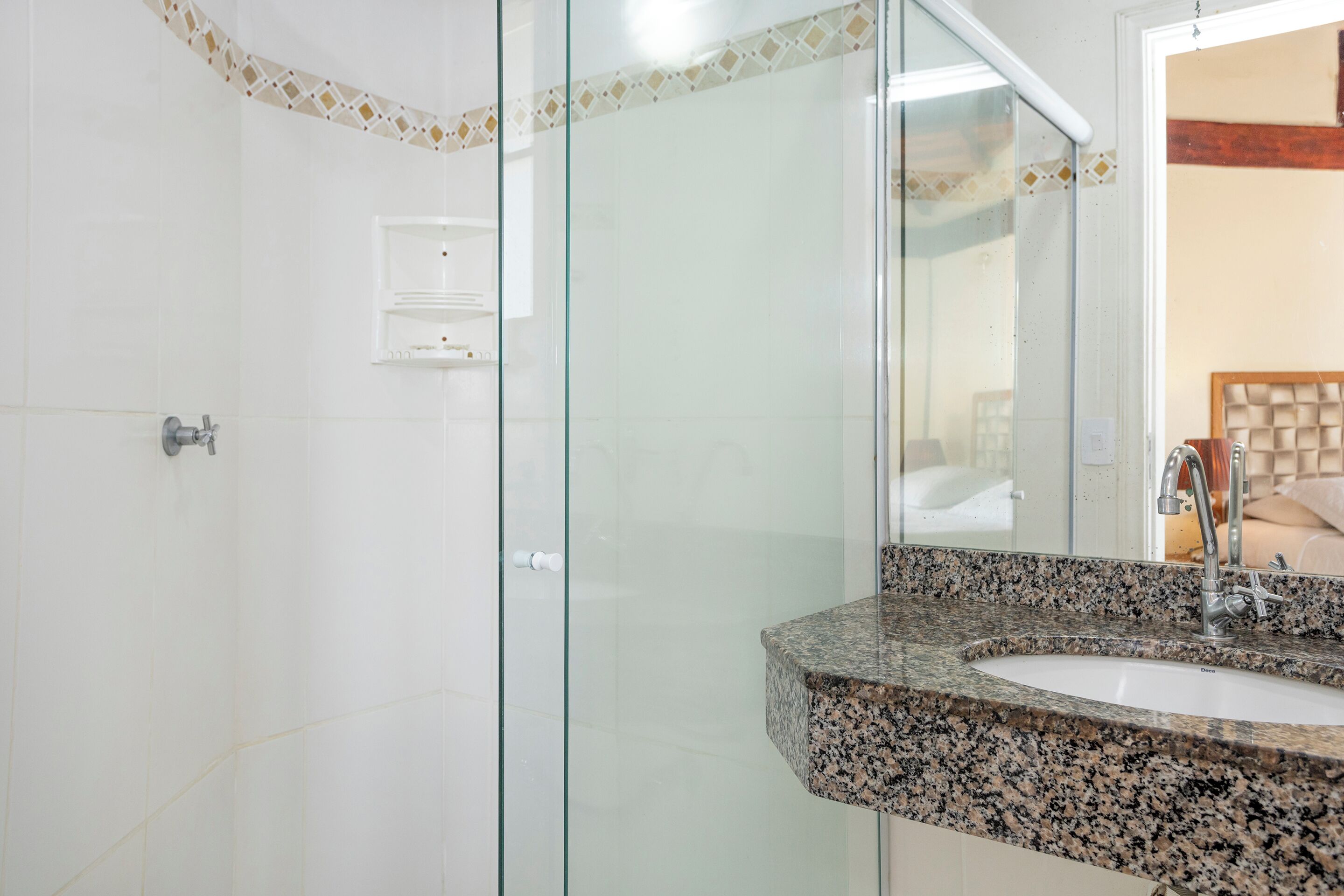 Un baño con lavamanos de encimera de granito, cabina de ducha de vidrio y pared cubierta de azulejos blancos.