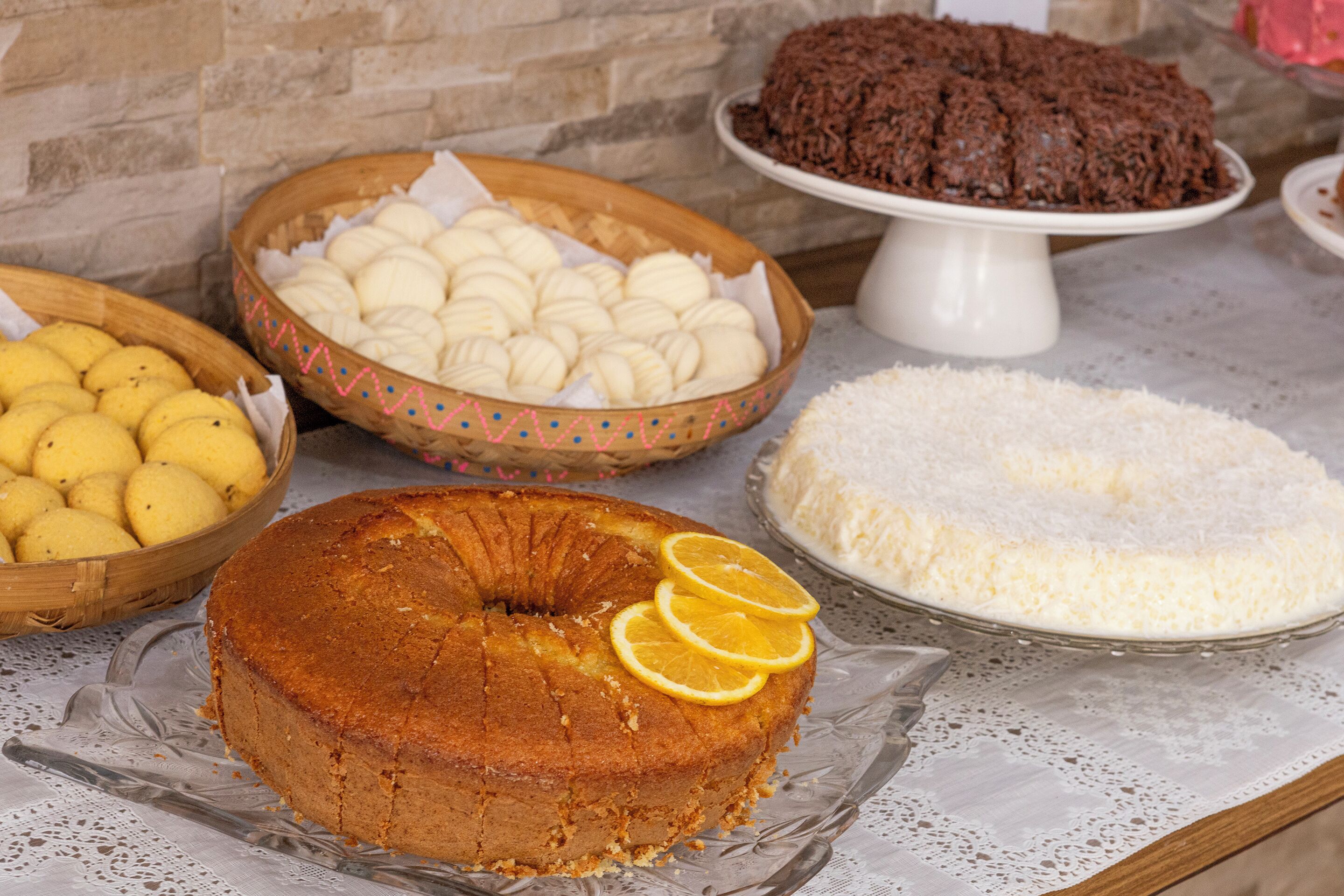 A variety of desserts including a bundt cake, lemon tarts, and a chocolate cake on a table with a lace tablecloth.