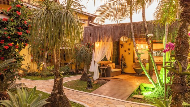 A tropical garden with a stone pathway leading to a seating area with a thatched roof and a sofa. - Pousada Bonns Ventos (Ilhabela)