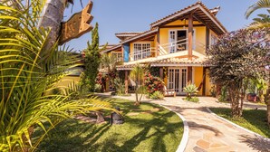 A two-story house with a stone pathway leading to the entrance, surrounded by lush greenery and tropical plants. - Pousada Bonns Ventos (Ilhabela)
