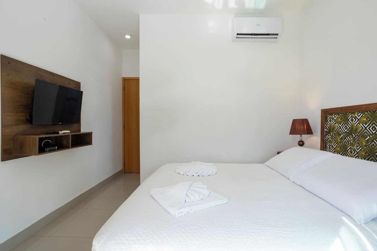 A hotel room with a bed, a television mounted on a wooden shelf, a wall-mounted air conditioner, and a patterned headboard.