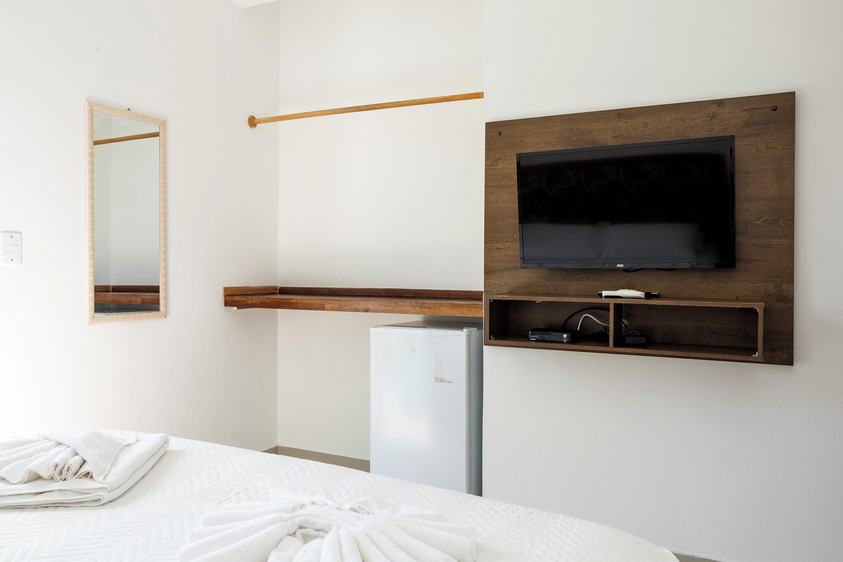 A hotel room with a bed, a TV mounted on a wooden panel, a mirror, and a refrigerator.