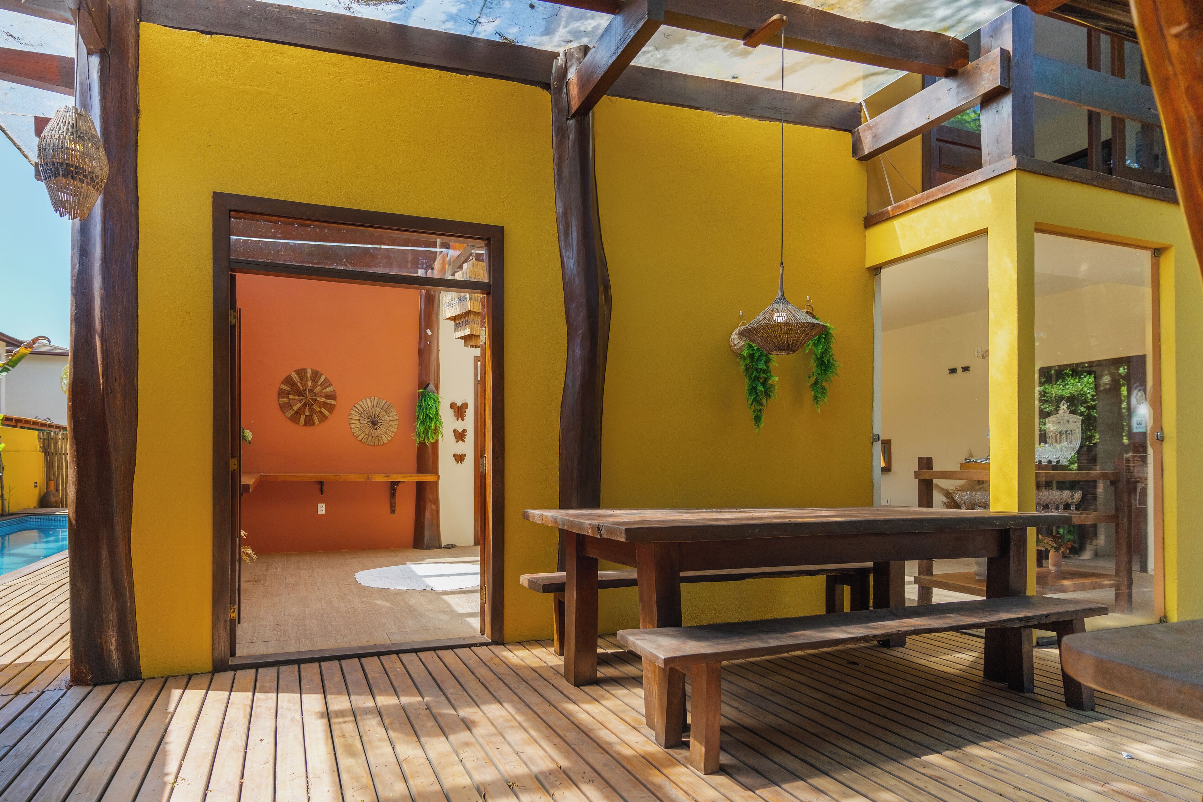A wooden deck with a bench and a table, a yellow wall, and a door leading to an interior space with a view of a pool.