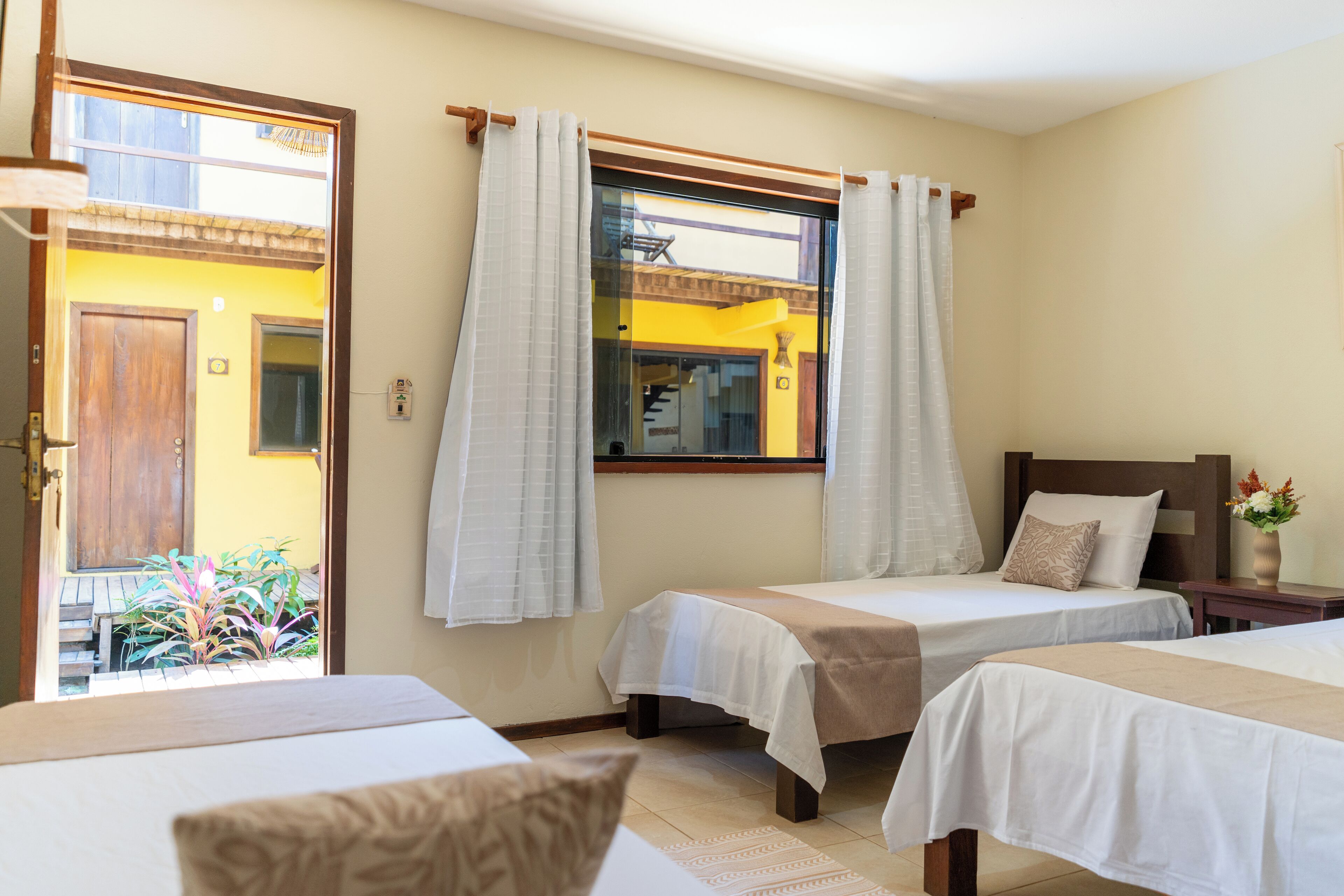A hotel room with two beds, a wooden door, a window with curtains, and a view of a yellow building outside.
