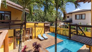 Zona de piscina con terraza de madera y sillones de descanso, rodeada de paredes amarillas y palmeras.
