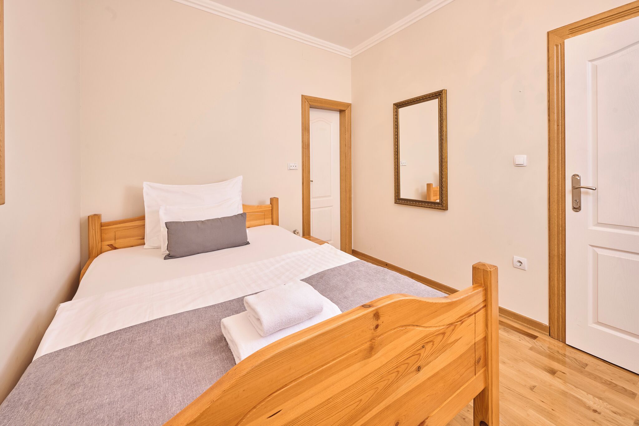 A neatly made bed with a wooden headboard, a single pillow, and a folded towel. There is a mirror on the wall and a door to the right.