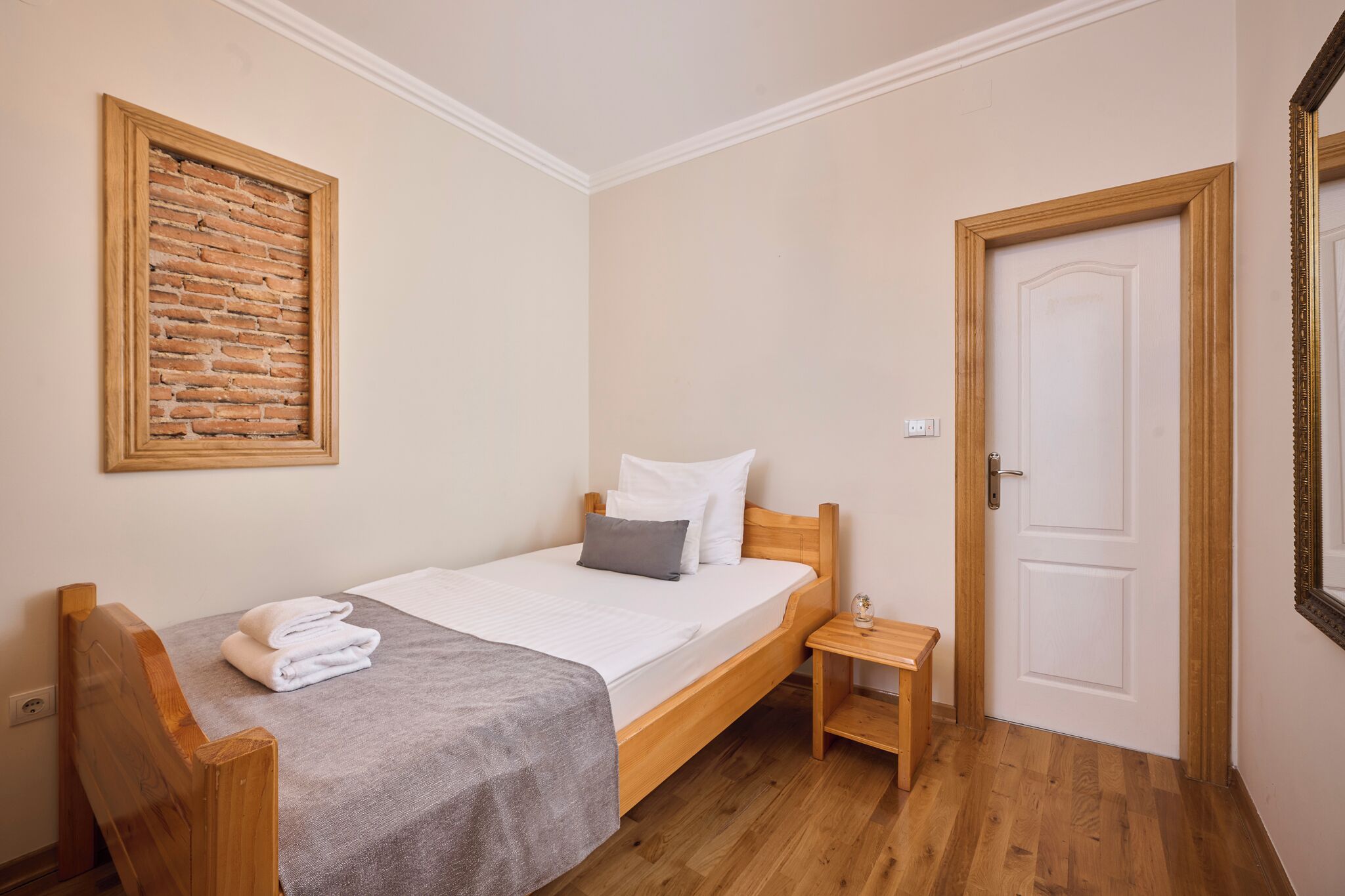 A single bed with a wooden headboard, a small bedside table, a framed picture on the wall, and a door with a mirror.