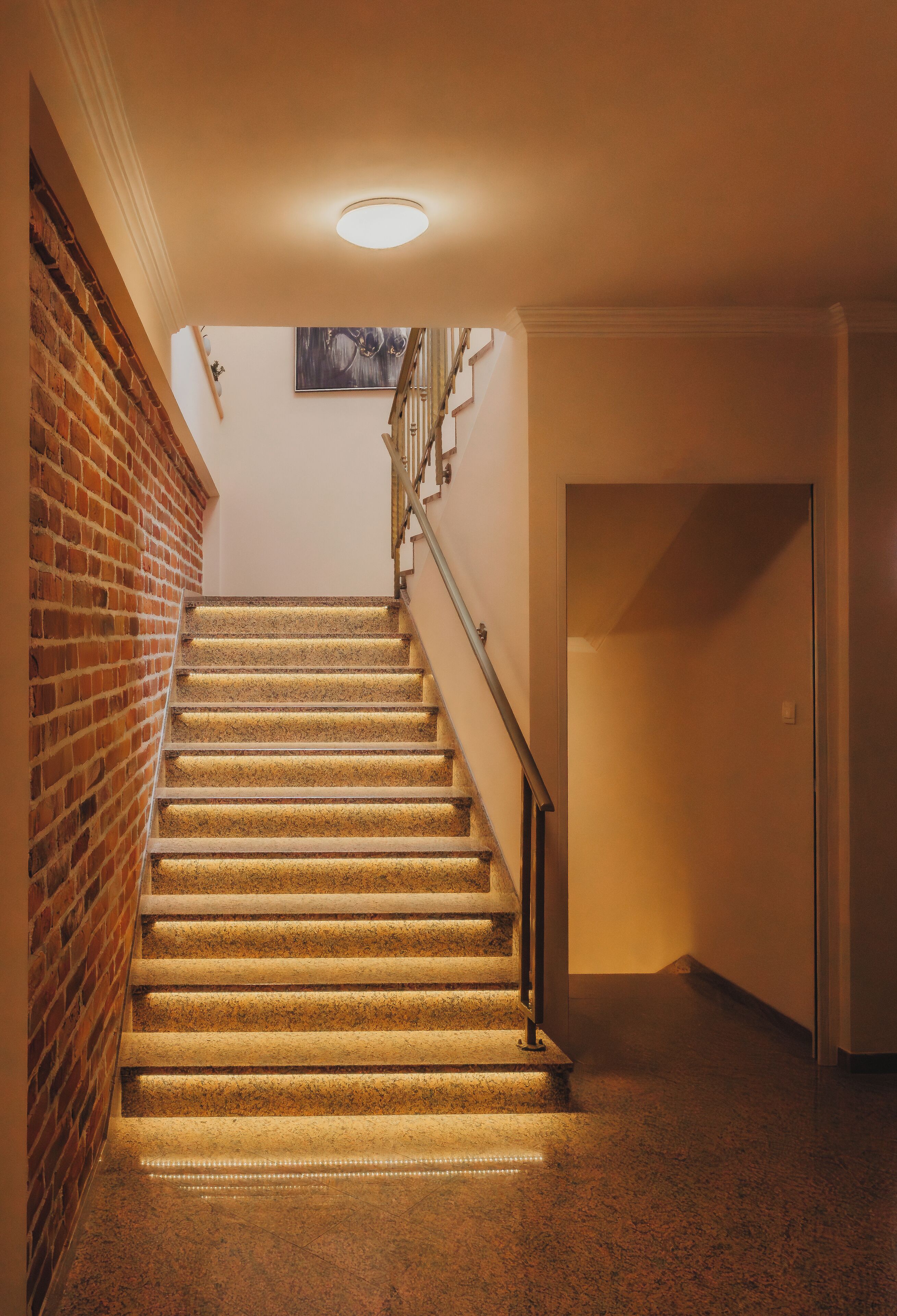Un escalier recouvert de moquette, doté d’une rampe, avec un mur en briques d’un côté et un mur uni de l’autre.