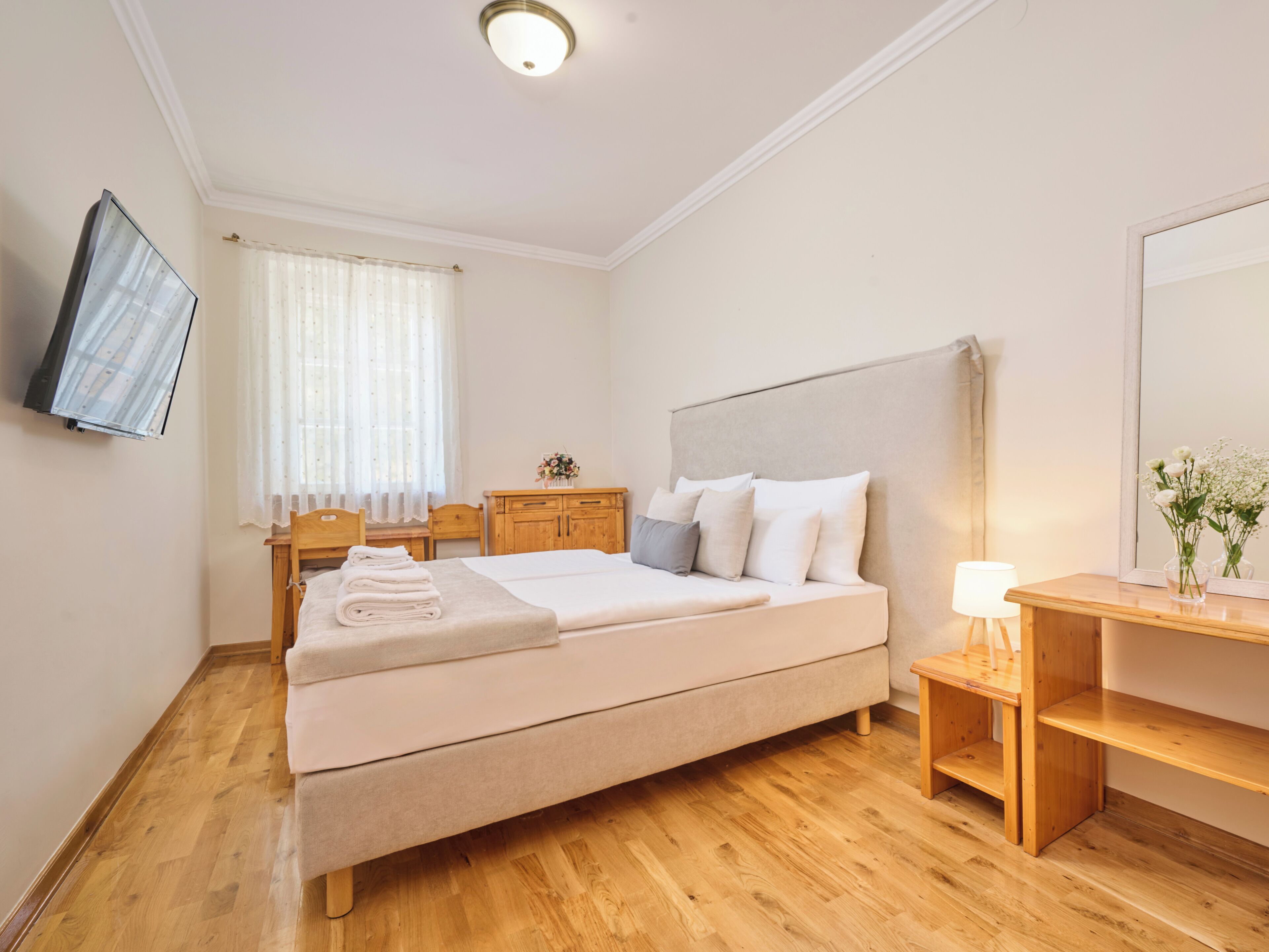 Habitación de hotel con cama, mesita de noche, lámpara, mesita auxiliar de madera y televisor montado en la pared.