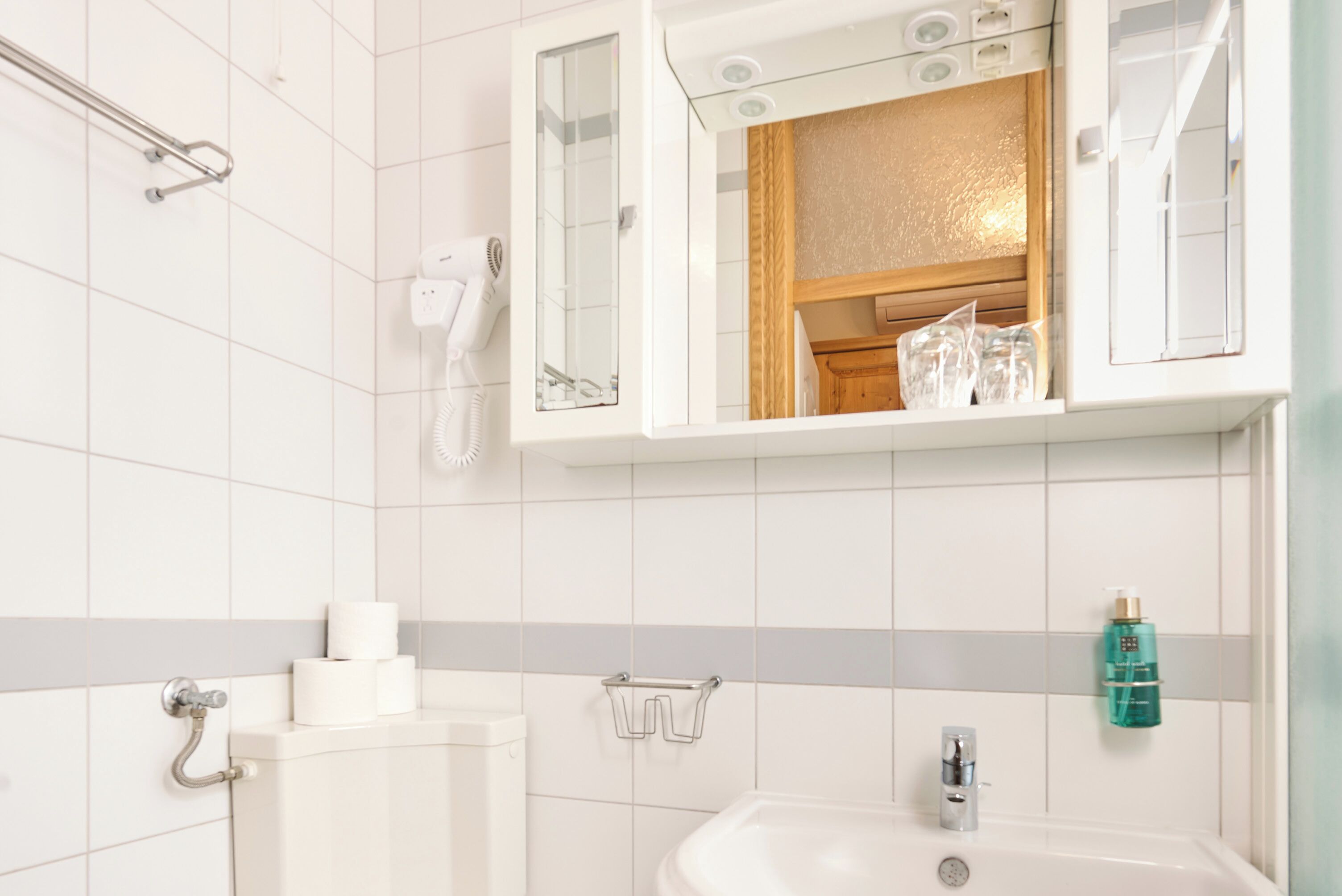 A bathroom with white tiles, a sink, a mirror, a towel rack, and a hand dryer.