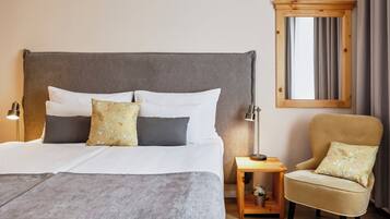 Una cama bien hecha con ropa de cama blanca y gris, una mesita de noche de madera con una lámpara, un sillón con cojines estampados, y una ventana con cortinas.