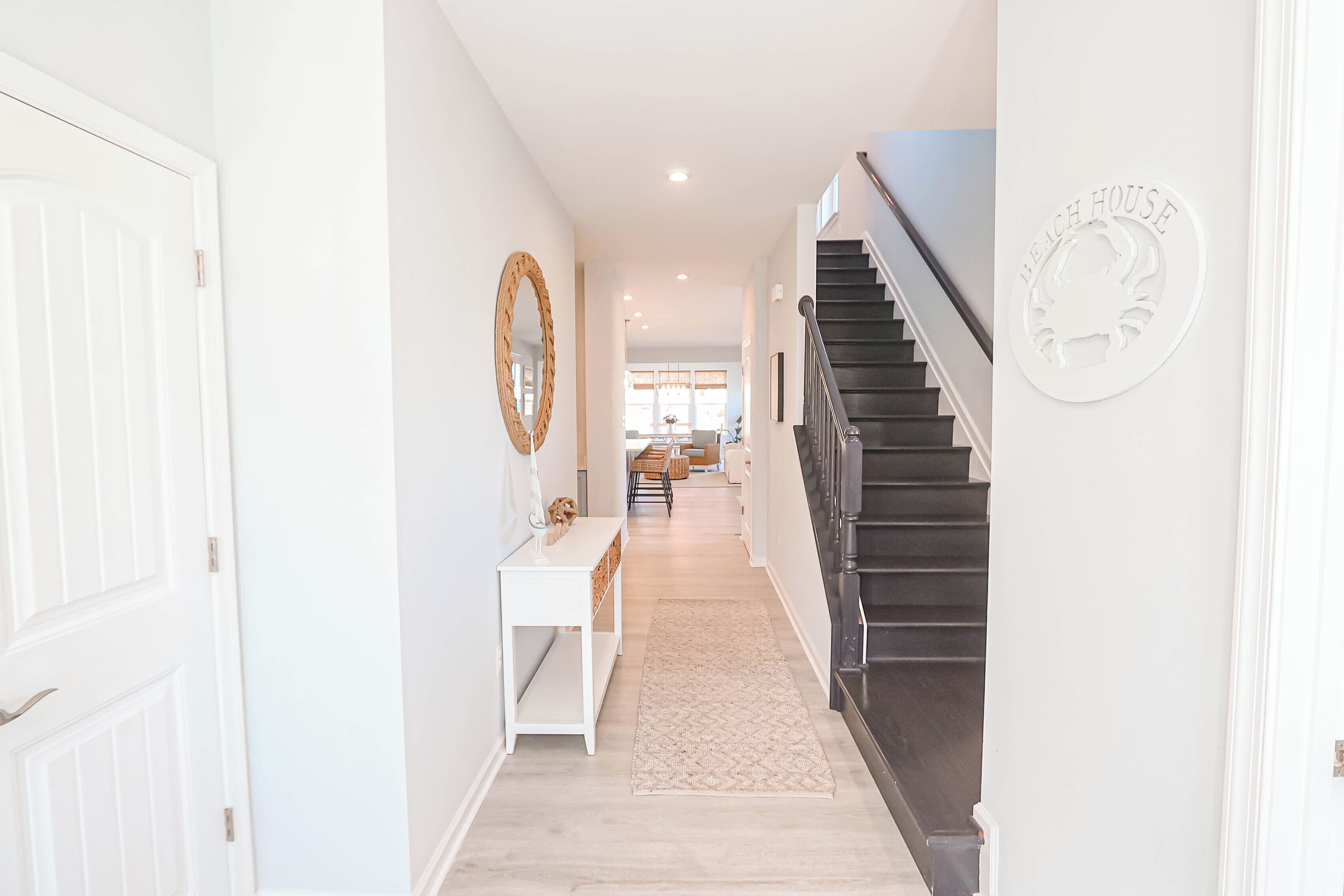 A hallway with a white door, a white console table, a patterned rug, and a staircase with a 'Beach House' sign.