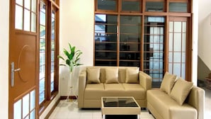 A spacious living room with a beige sofa, a glass coffee table, and large windows with wooden frames. - SF Homey Puri Indah (Cipanas)