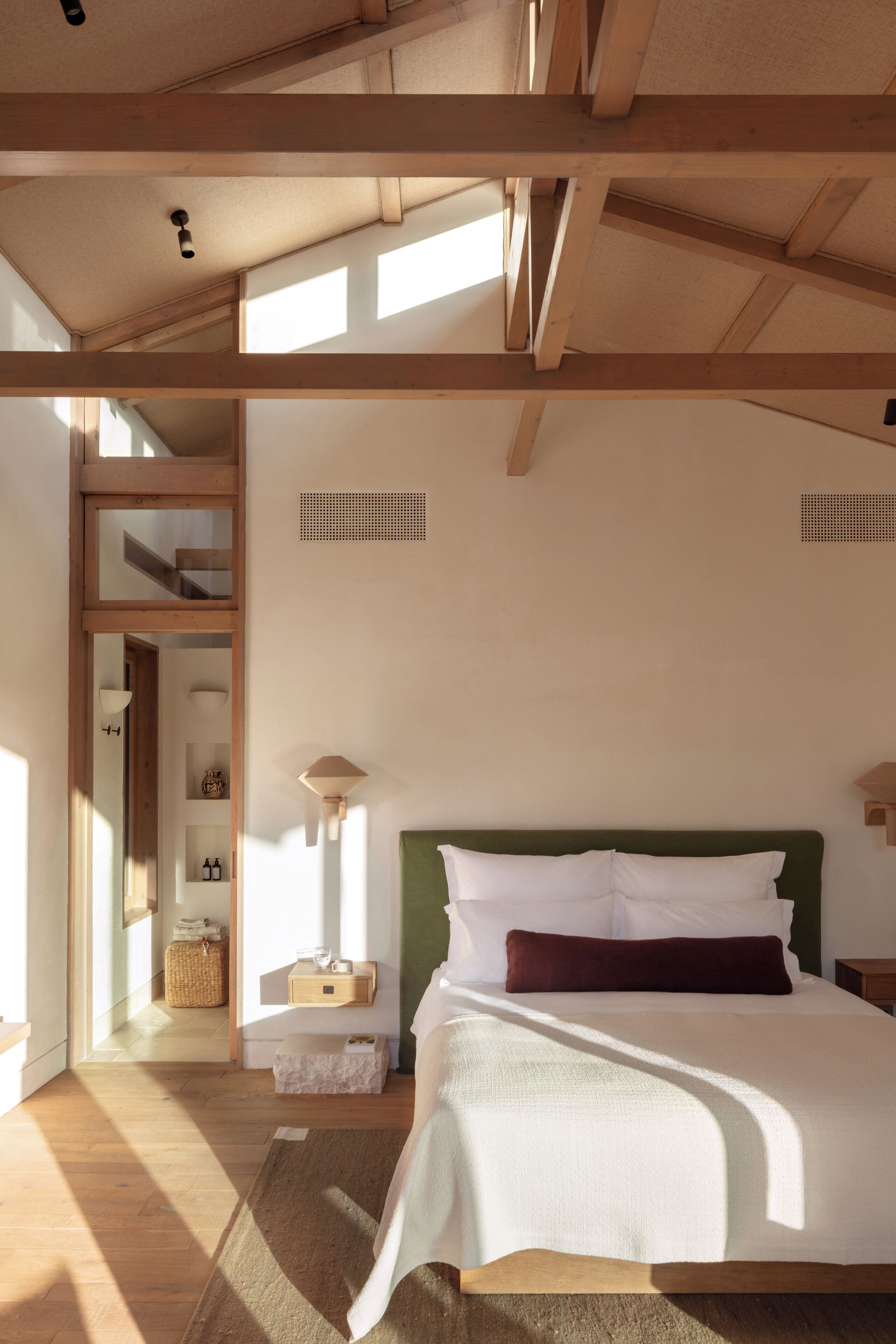 A bedroom with a large bed, wooden beams, a green headboard, and a view of a hallway.