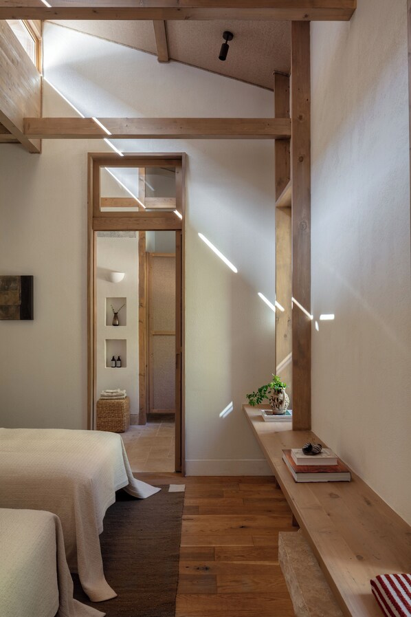 A room with wooden beams, a bed, a bench, and a small table with books and a plant.