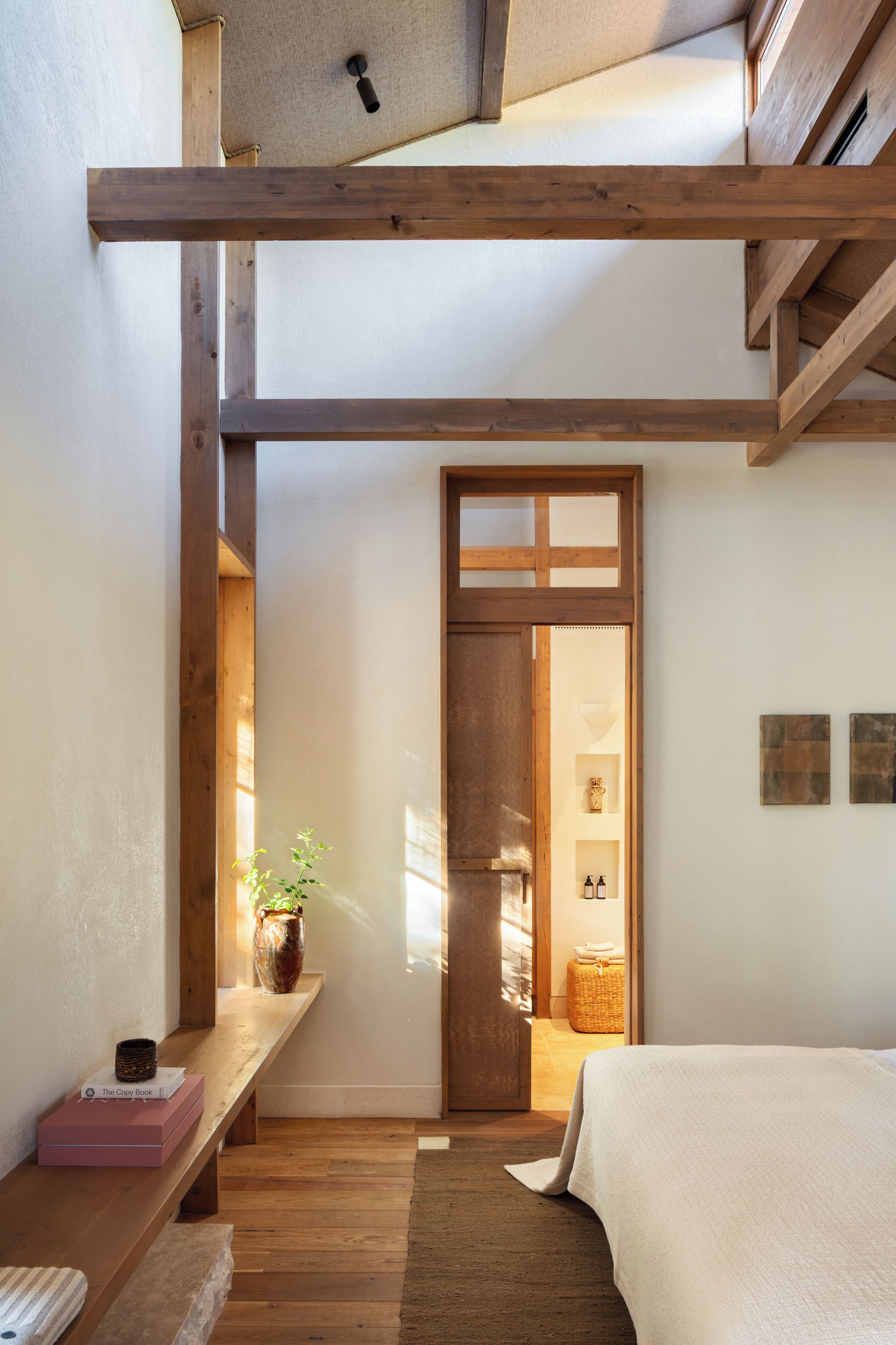 Un dormitorio con techo de madera, una cama, un banco con libros, un jarrón y una puerta que da a otra habitación.