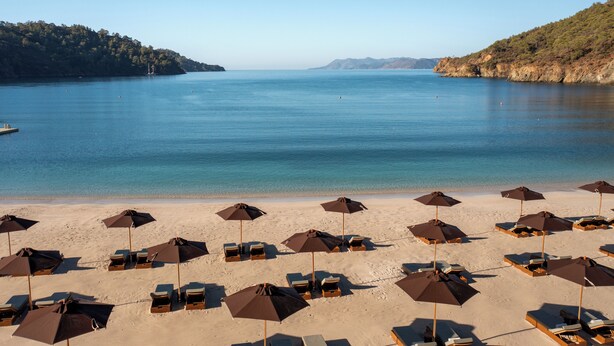 A beach with rows of sun loungers and umbrellas, a calm sea, and a hilly landscape in the background. - Ahama - Relais & Châteaux (Fethiye)