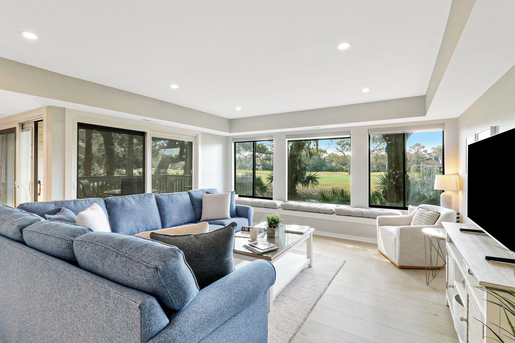 A spacious living room with a large sectional sofa, a coffee table, and a TV. The room has large windows offering a view of a garden.