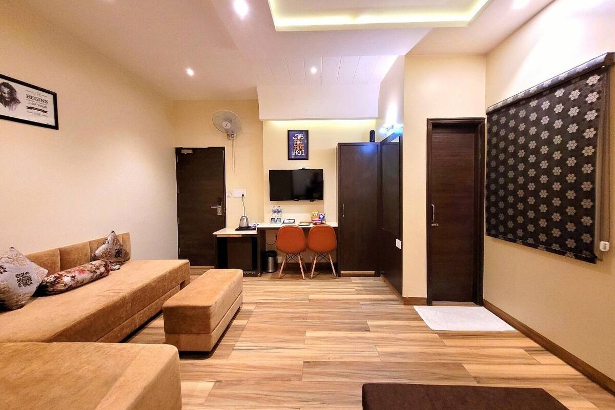 Ein modernes Wohnzimmer mit einer Couch, einem Couchtisch und einem an der Wand montierten Fernseher. Es gibt einen Schreibtisch mit zwei Stühlen und einen kleinen Tisch mit einem Laptop. Der Raum hat Holzböden und einen gemusterten Vorhang am Fenster.