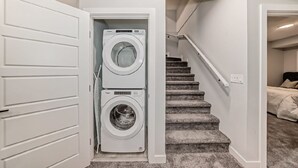 A laundry room with two stacked front-loading washing machines, a carpeted staircase, and a bedroom with a bed and pillows.