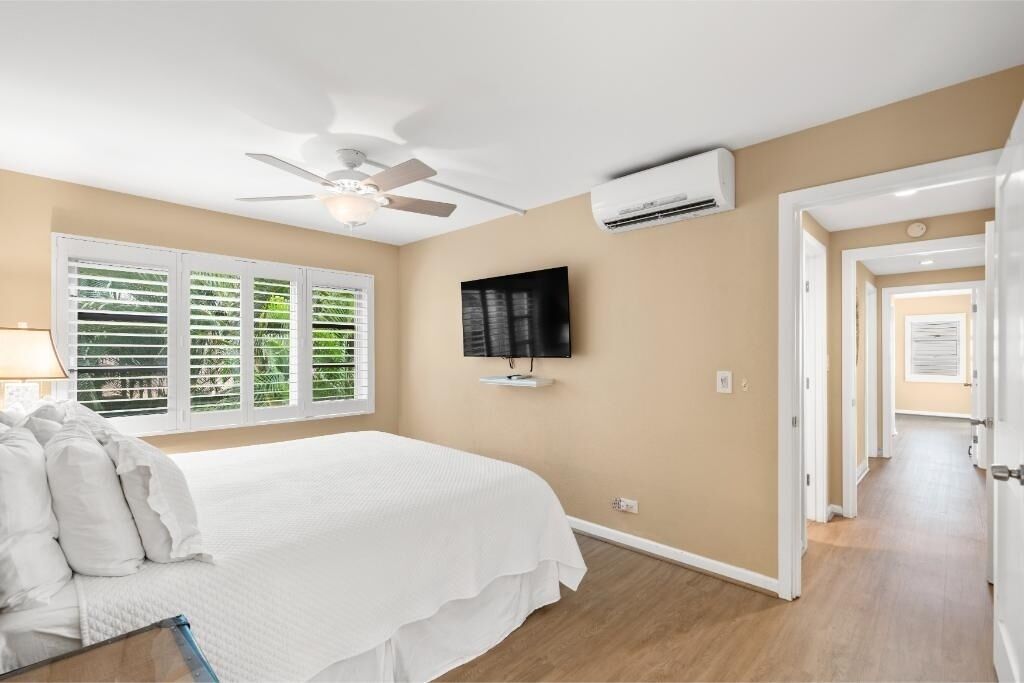 A bedroom with a bed, a ceiling fan, an air conditioning unit, a TV, and a window with blinds.