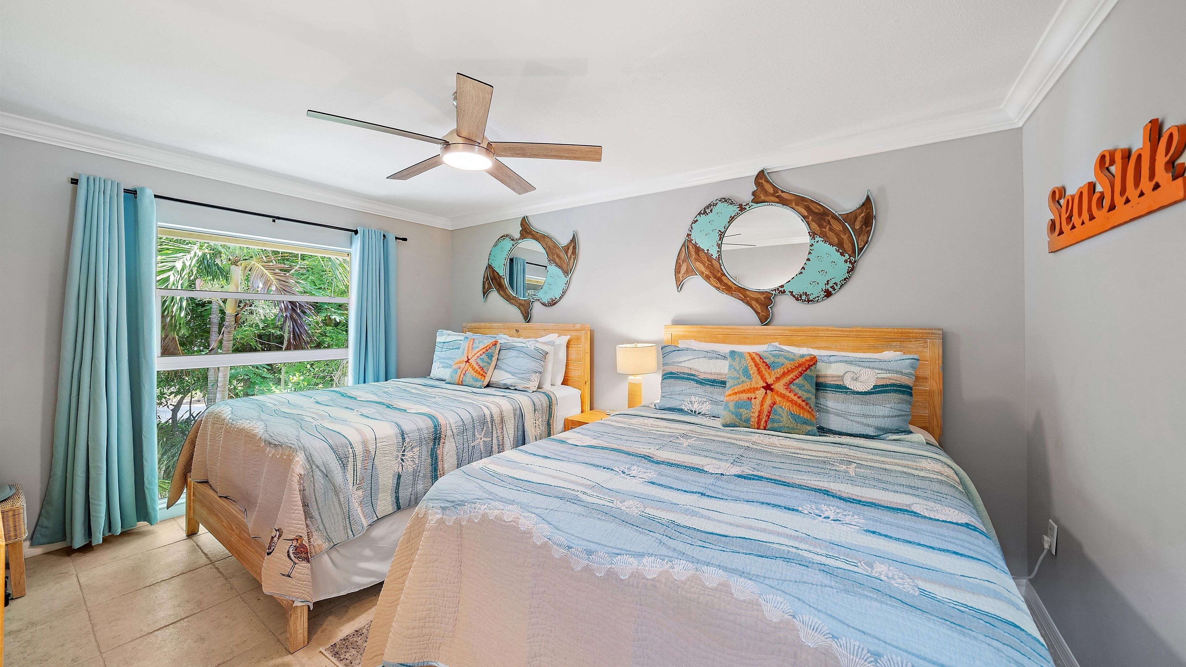 Una camera da letto con due letti, un ventilatore a soffitto, un'ampia finestra con tende e un elemento decorativo a parete.