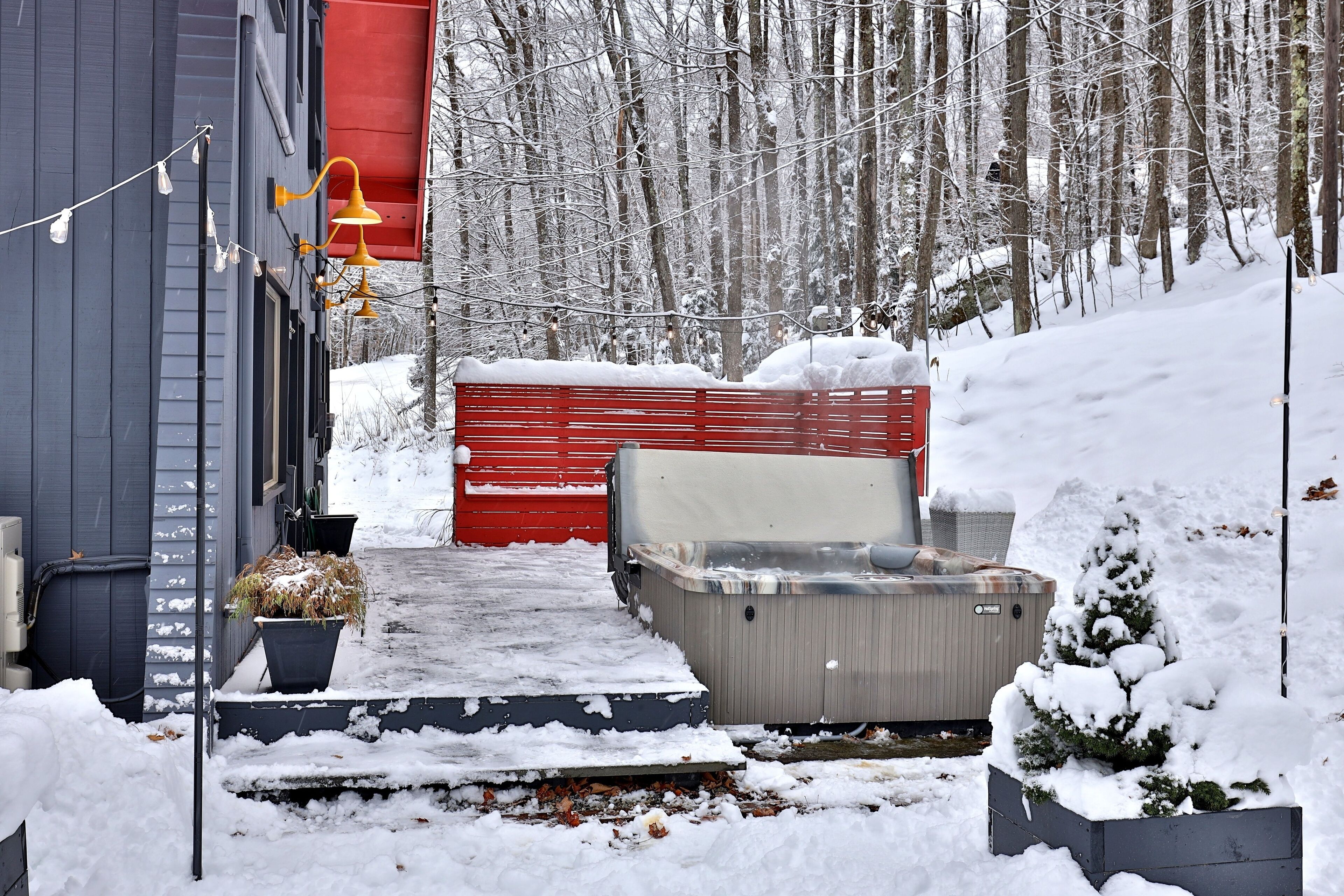 A snow-covered outdoor area with a red shed, a hot tub, and a small tree in a planter.