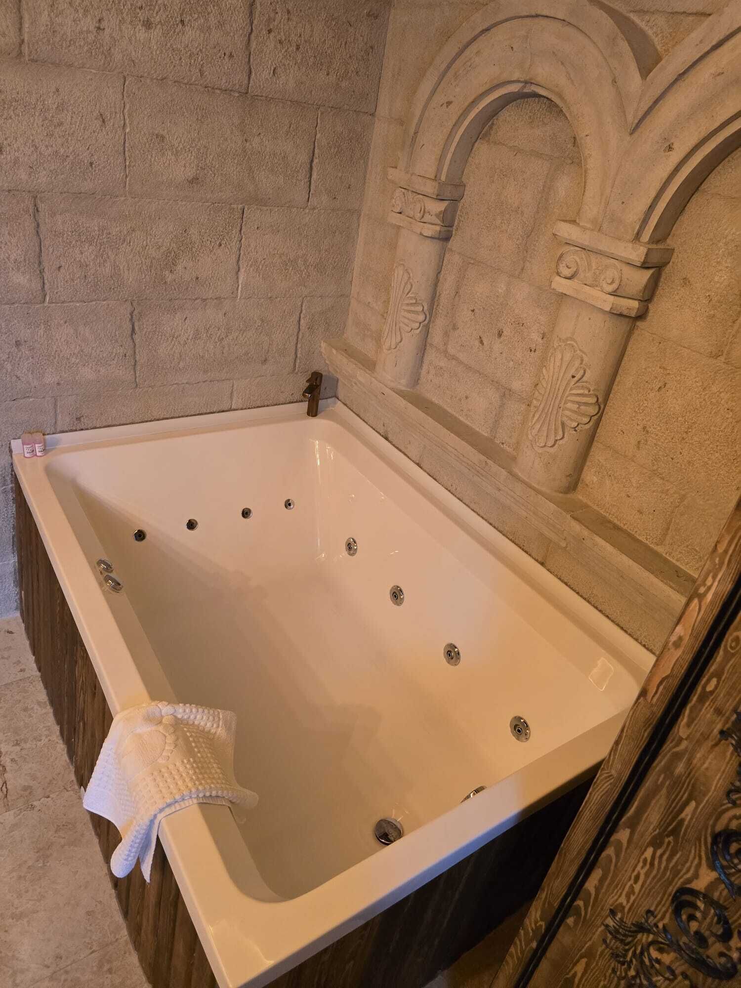 A large, empty bathtub with a white interior, set against a stone wall with decorative arches.