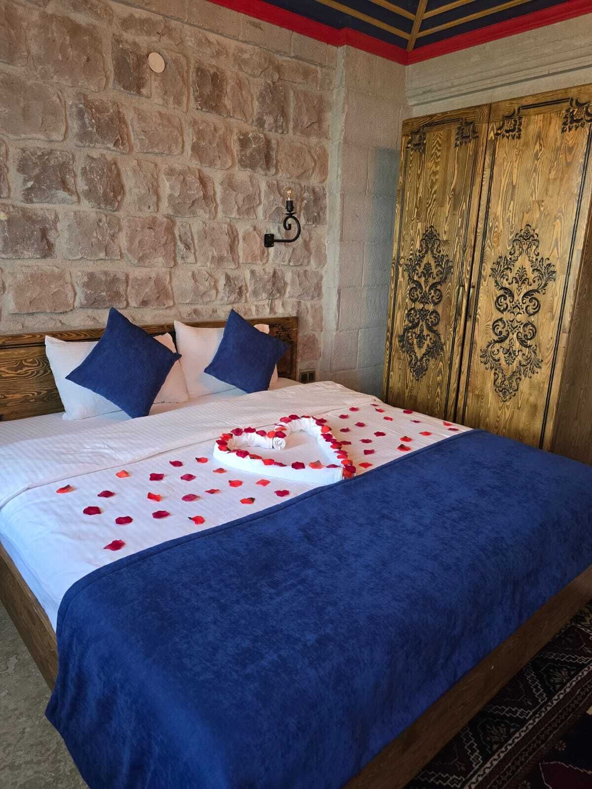 A bed with a heart-shaped towel and rose petals, a stone wall, and a wooden wardrobe with intricate carvings.