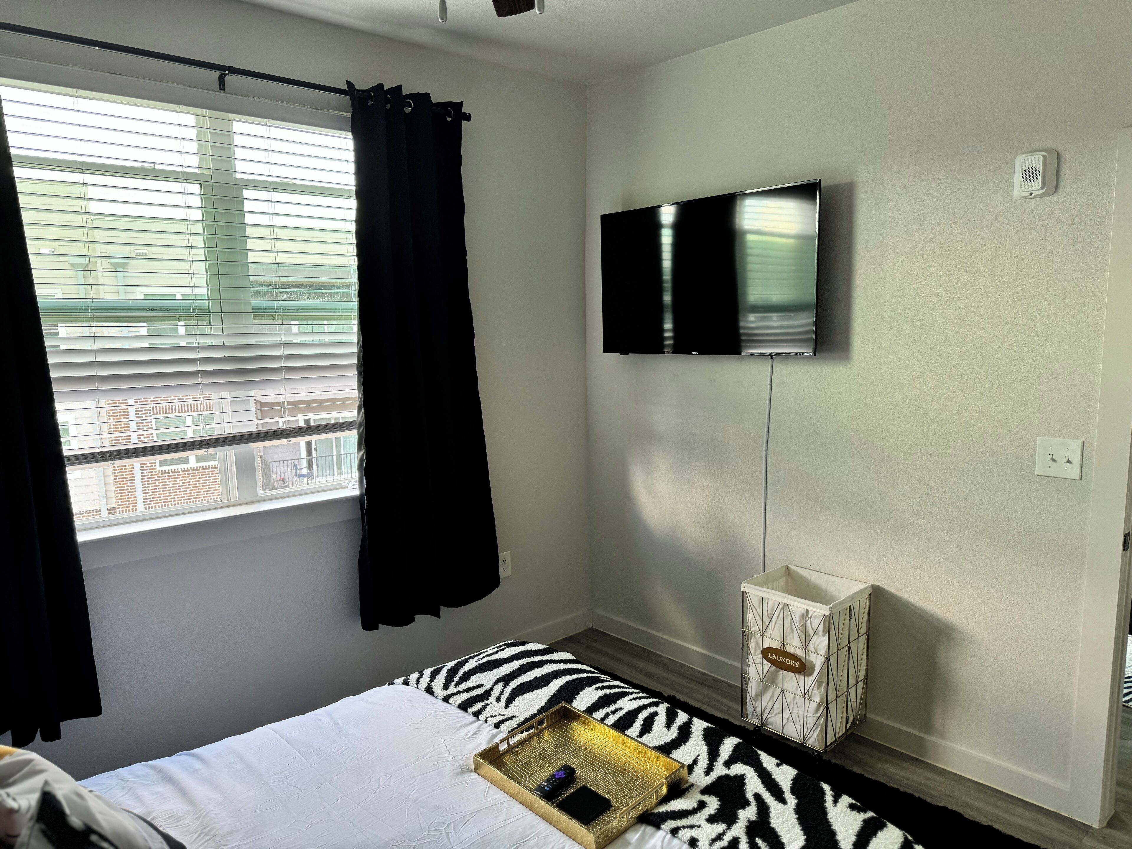 Une chambre à coucher avec un lit, un téléviseur fixé au mur, une fenêtre avec des stores et un panier à linge.