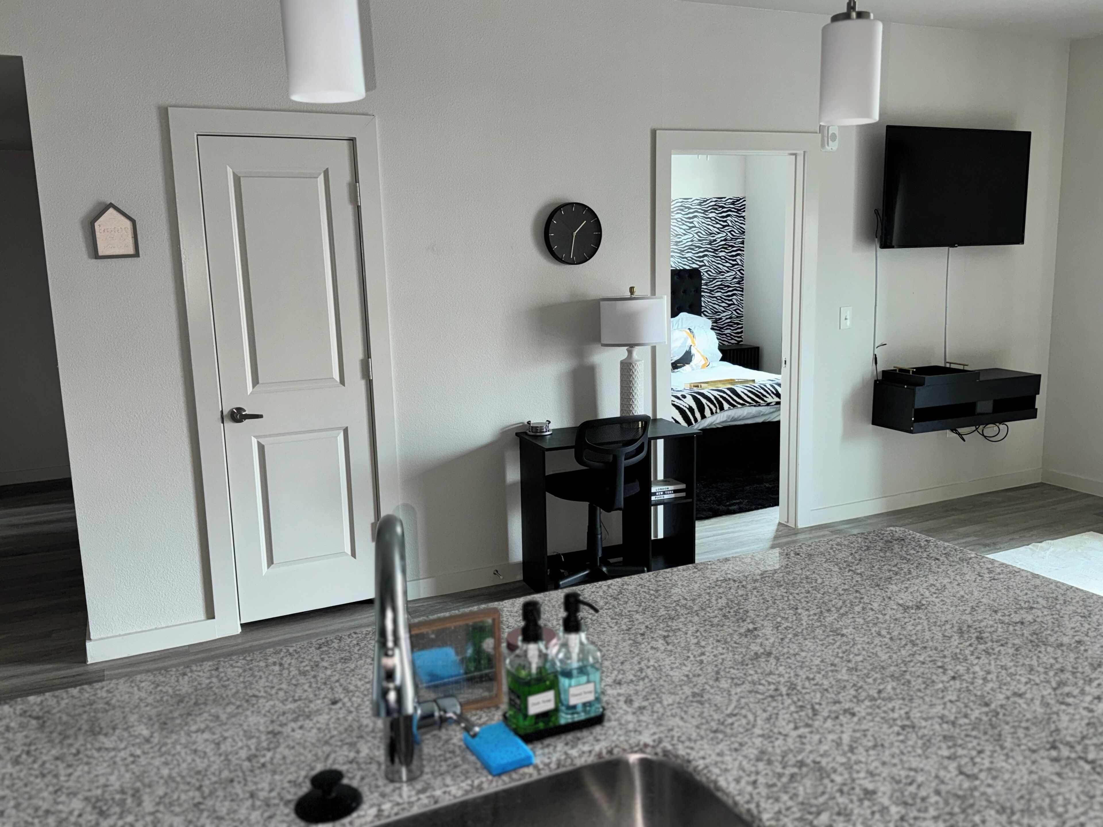 A modern hotel room with a kitchenette, a bed with zebra print bedding, a mounted TV, and a small desk with a chair.