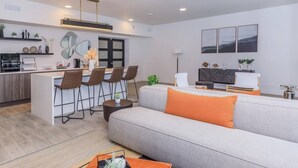 A modern living room with a grey sofa, orange cushions, a dining area with bar stools, a kitchen with a white countertop, and a large mirror on the wall. - Your Cozy & Captivating Condo! (Houston)