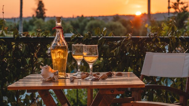 Une table en bois avec une bouteille et deux verres, entourée de coquillages, le tout sur fond de coucher de soleil.