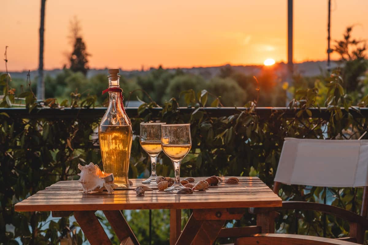 Ein Holztisch mit einer Flasche und zwei Gläsern, umgeben von Muscheln, vor einem Sonnenuntergang.
