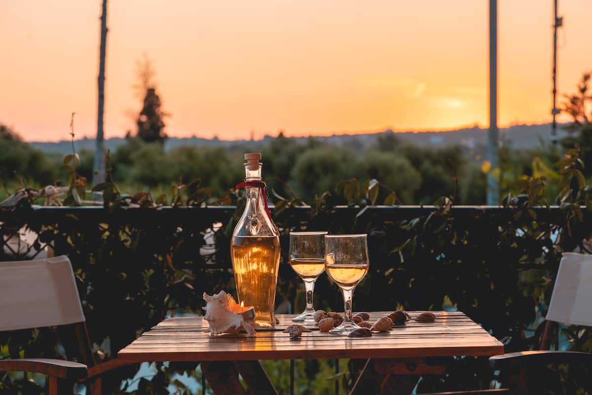 Ein Tisch im Freien mit einer Flasche und zwei Gläsern, umgeben von Grünpflanzen und einem Sonnenuntergang im Hintergrund.