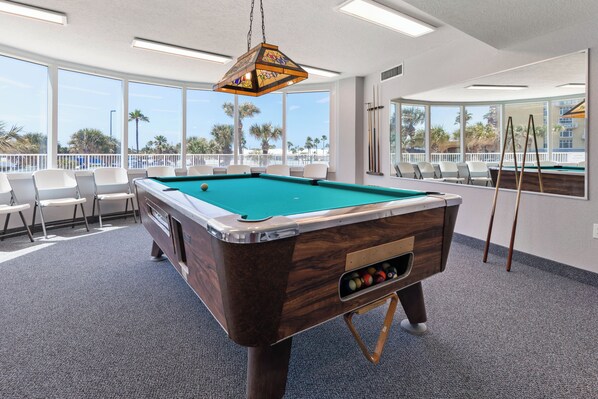 A pool table in a room with large windows, chairs, and a pool table with a wooden finish.