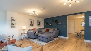 A modern living room with a sectional sofa, a small side table, and a lamp. There are framed pictures on the wall and a ceiling light fixture.