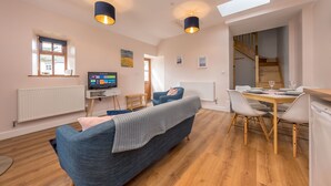 A modern living room with a sofa, a dining table with chairs, a TV, and a staircase leading to another level. - Two bedroom barn conversion on Anglesey | Bwthyn-Cerrig (Llannerch-y-medd)